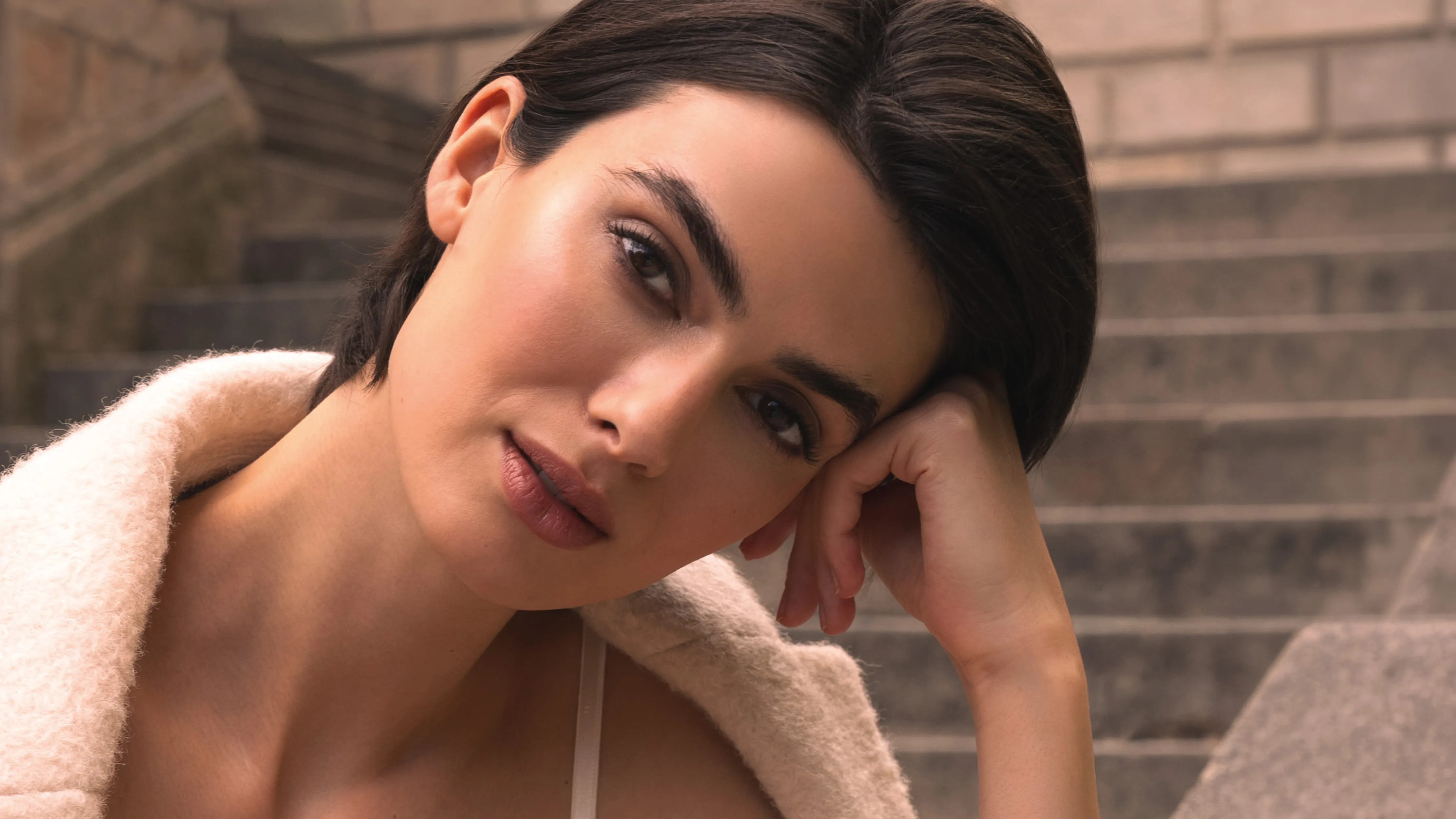 Beauty-Porträt einer Frau mit kurzem dunklem Haar, die ihren Kopf auf die Hand stützt und vor einer Steintreppe in die Kamera blickt.
