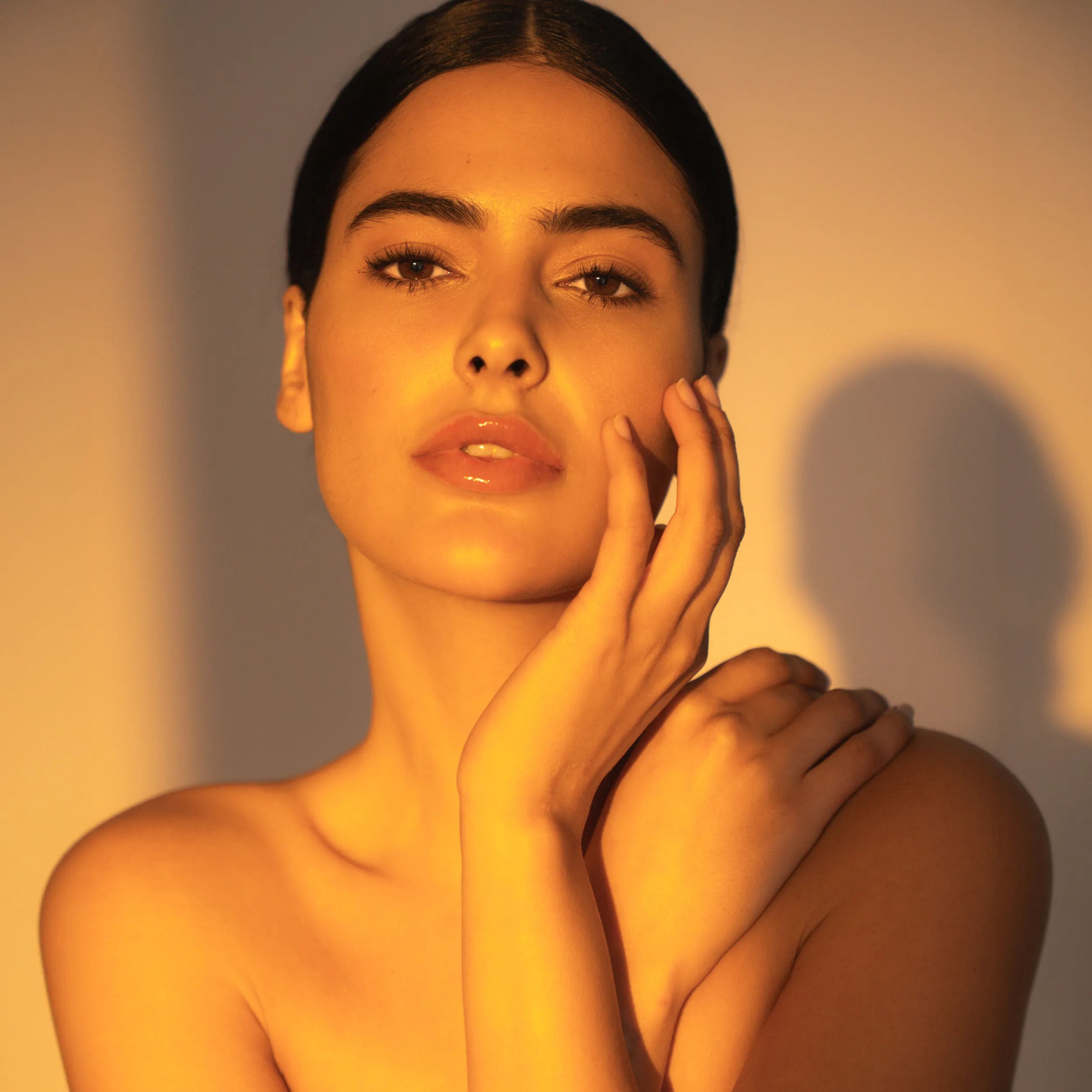 Beauty-Porträt eines Models mit zurückgebundenem Haar und natürlichem Make-up im warmen Sonnenlicht, das die Hand sanft an die Wange legt.