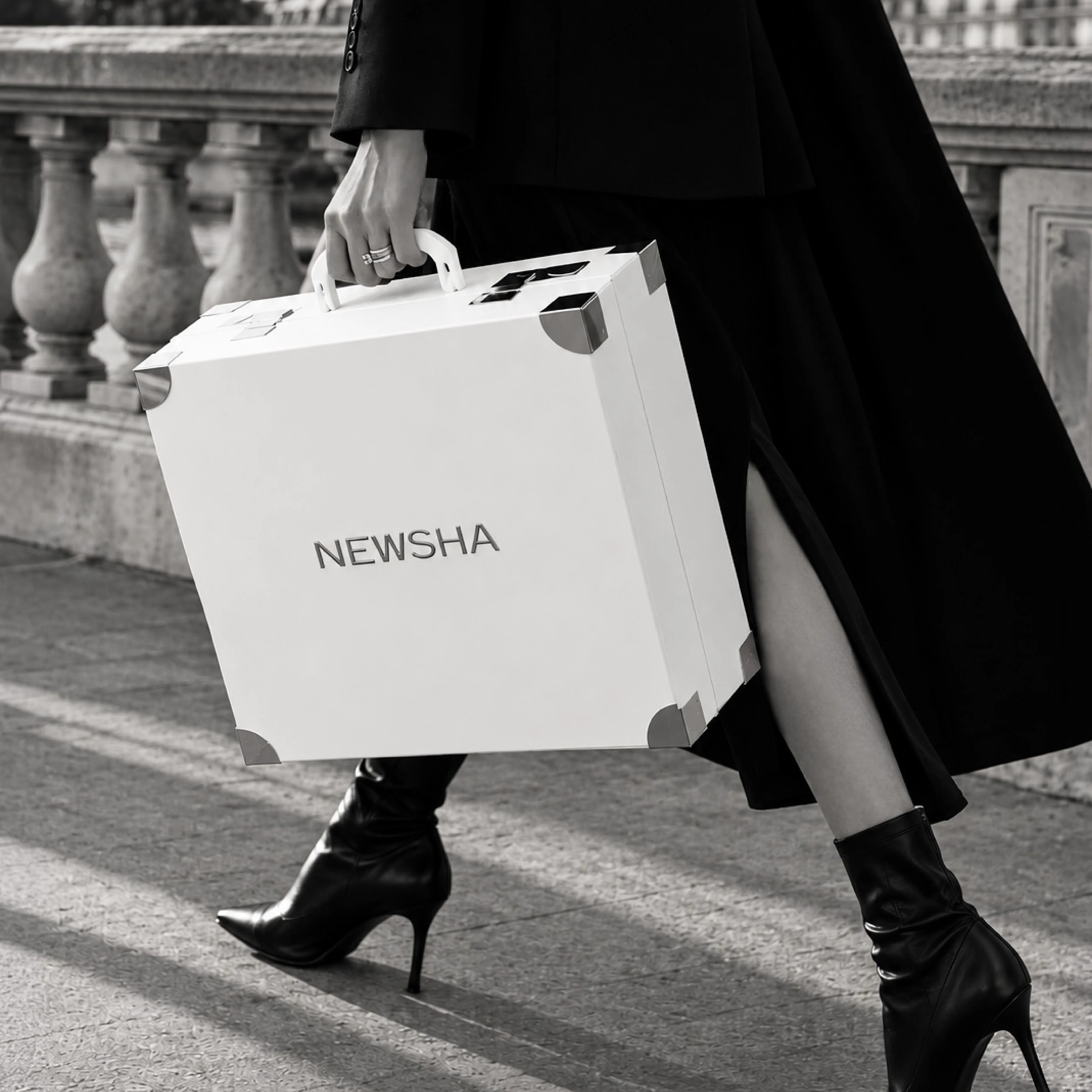 Elegant gekleideten Person in schwarzem Outfit und hohen Stiefeletten, die dem NEWSHA Adventskalender mit der Aufschrift &bdquo;NEWSHA&ldquo; tr&auml;gt. Im Hintergrund ist eine steinerne Balustrade zu sehen.