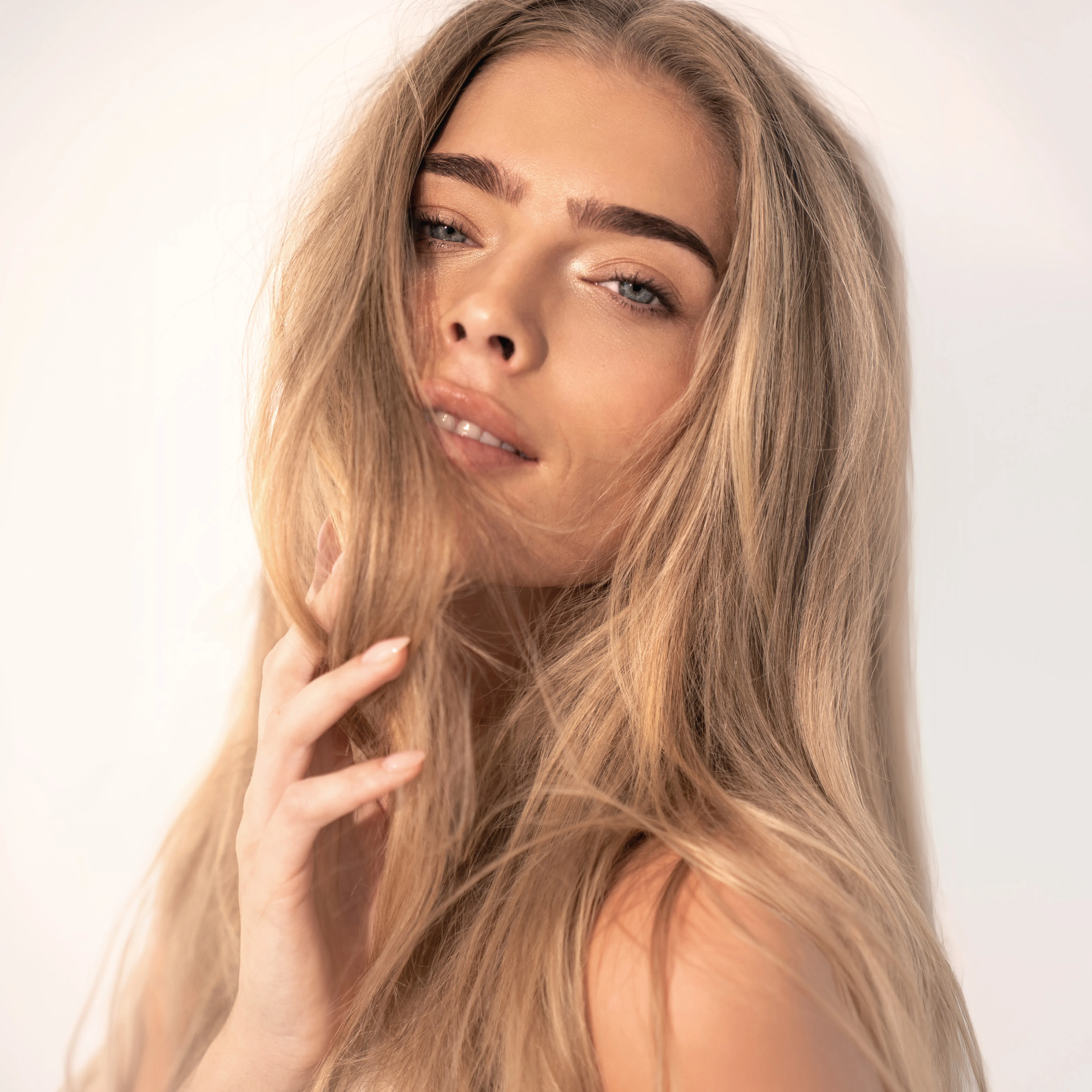Frau mit langen, glänzenden blonden Haaren und natürlichem Make-up vor hellem Hintergrund.