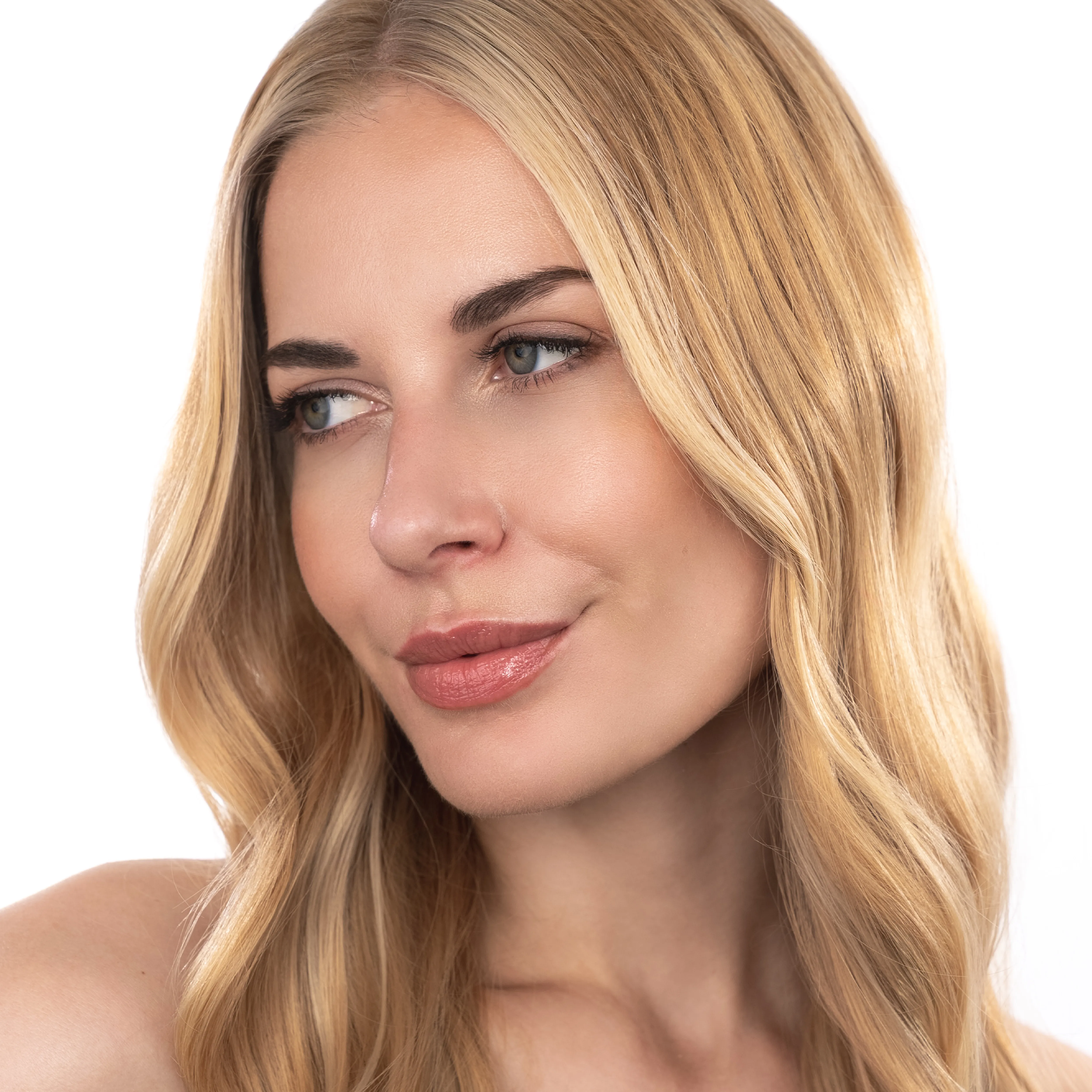 Blonde Frau mit gl&auml;nzenden, weichen Wellen und nat&uuml;rlichem Make-up blickt seitlich vor hellem Hintergrund
