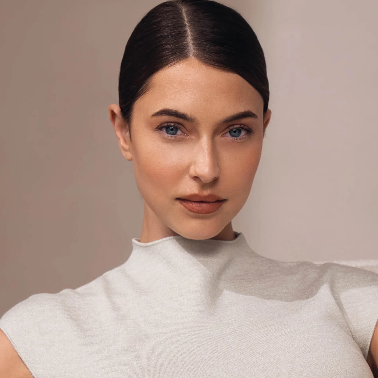 Portr&auml;t eines Models mit sleek bun, nat&uuml;rlichem Make-up und klarem Blick in die Kamera vor einem warmen, minimalistischen Studiohintergrund.