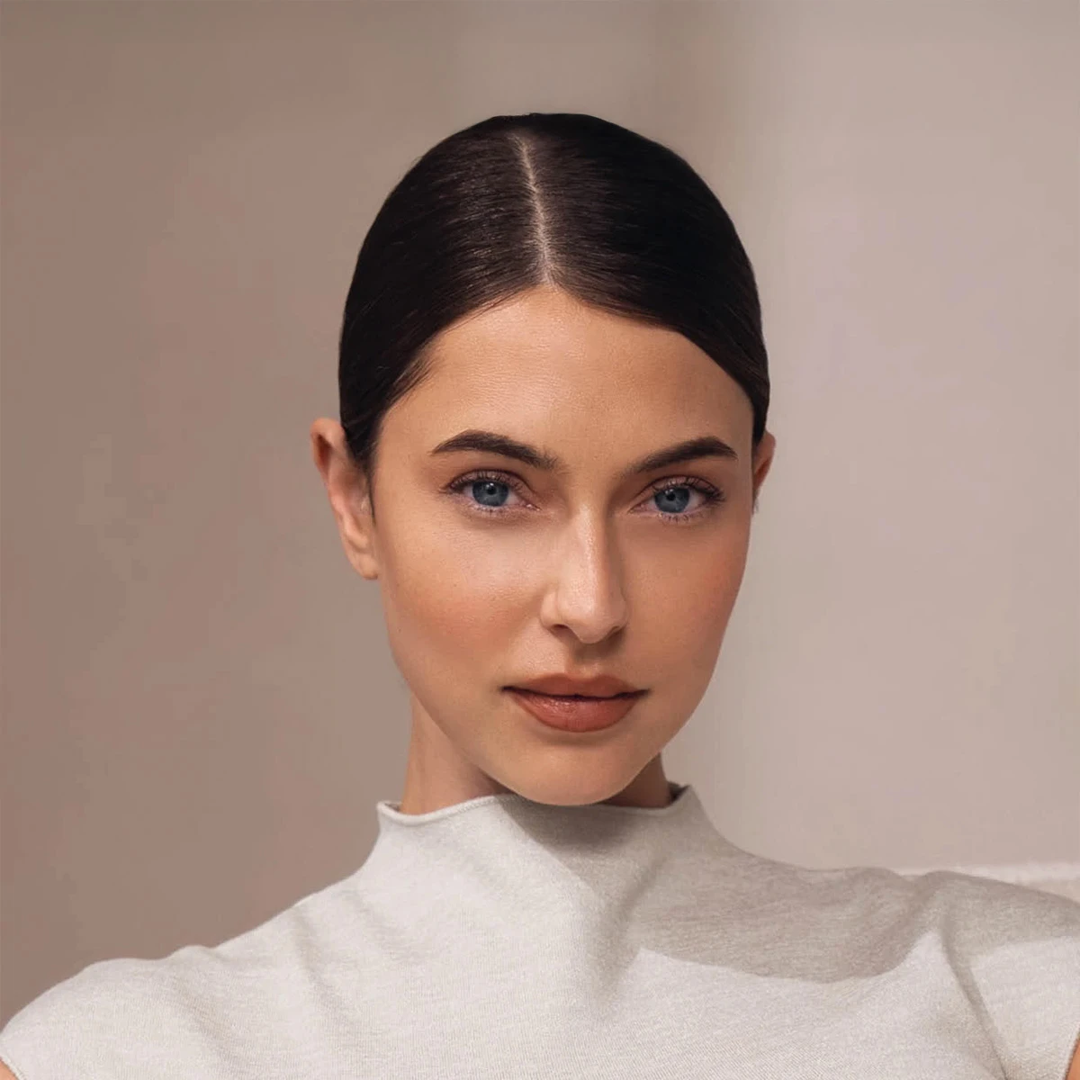 Portr&auml;t eines Models mit sleek bun, nat&uuml;rlichem Make-up und klarem Blick in die Kamera vor einem warmen, minimalistischen Studiohintergrund.
