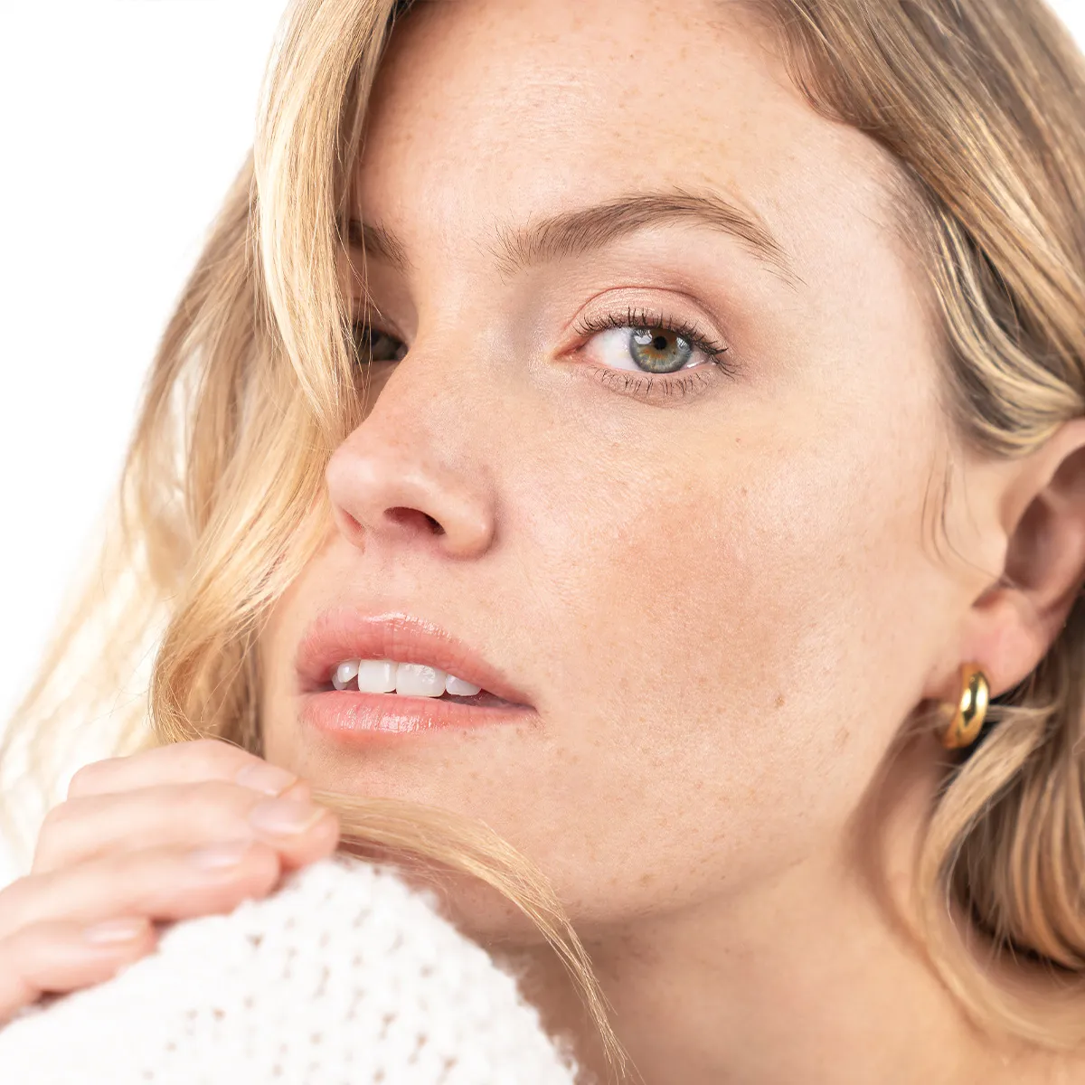 Nahaufnahme eines Models mit nat&uuml;rlicher Hautstruktur und Sommersprossen, sanft gewelltem blondem Haar und dezentem Beauty-Look vor hellem Hintergrund.