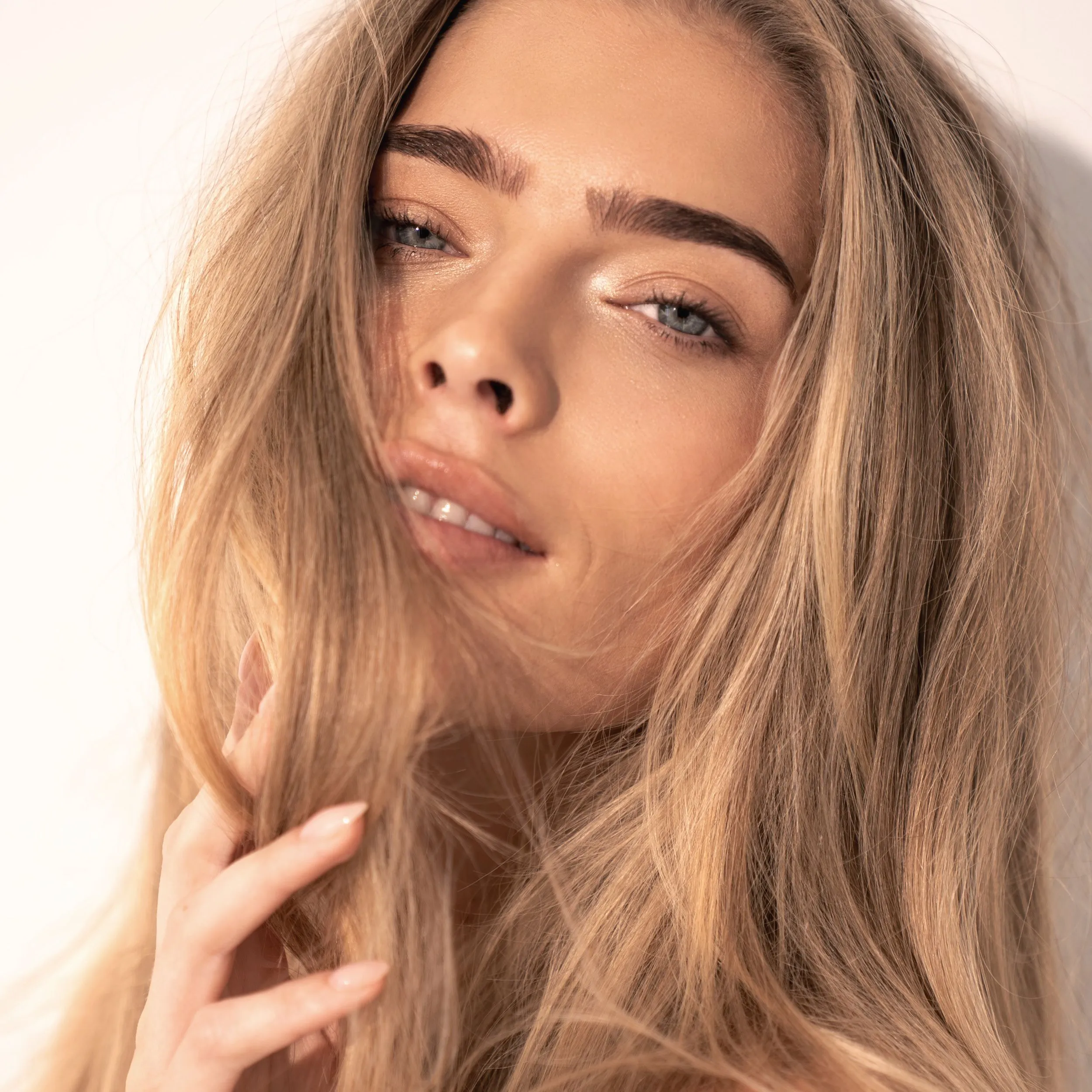 Nahaufnahme eines Models mit langem blondem Haar, definierten Augenbrauen und nat&uuml;rlichem Glow-Look vor hellem Hintergrund.
