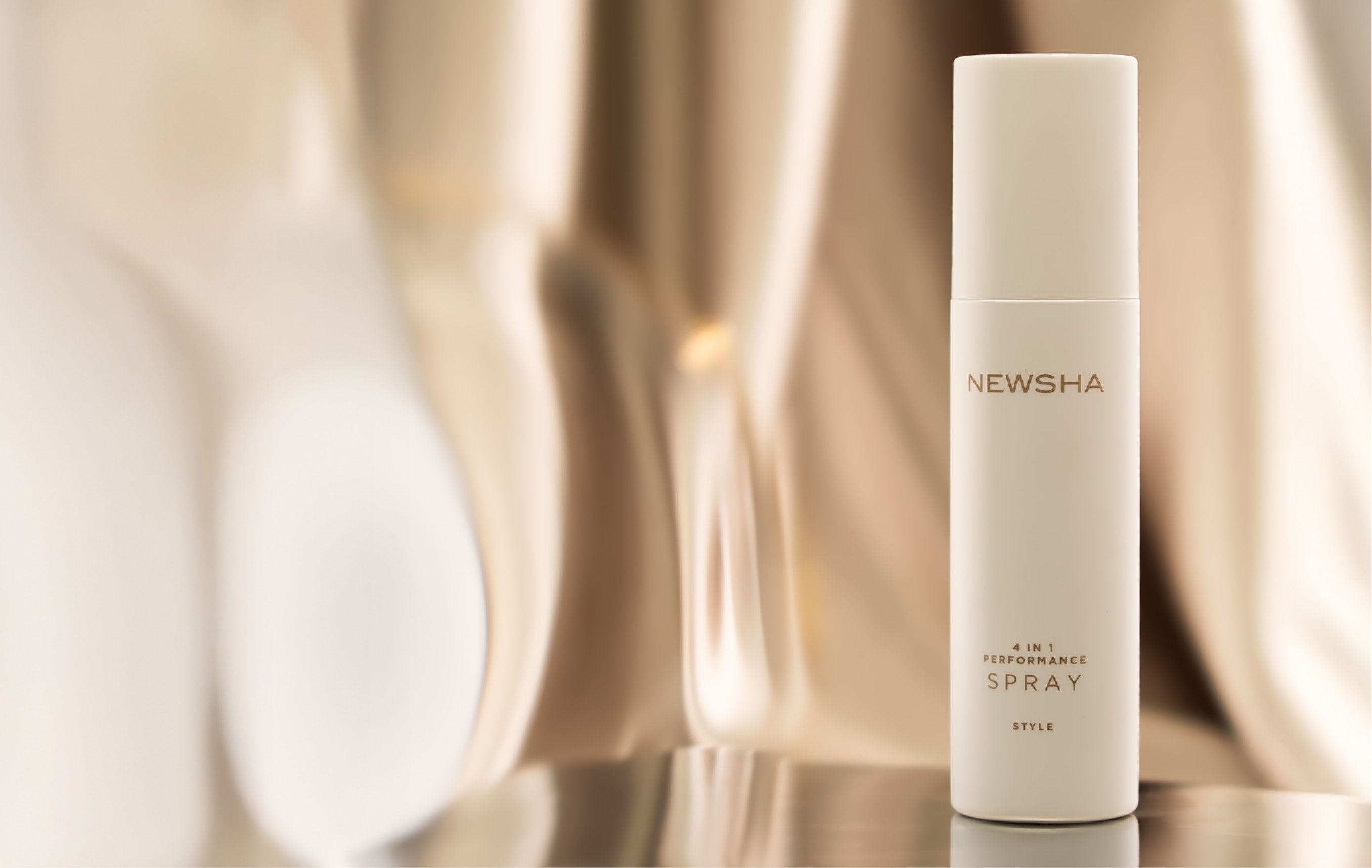 NEWSHA 4 in 1 Performance Spray als hochwertiger Packshot auf spiegelnder Oberfläche vor elegantem, warm beleuchtetem Hintergrund mit sanften Reflexionen.