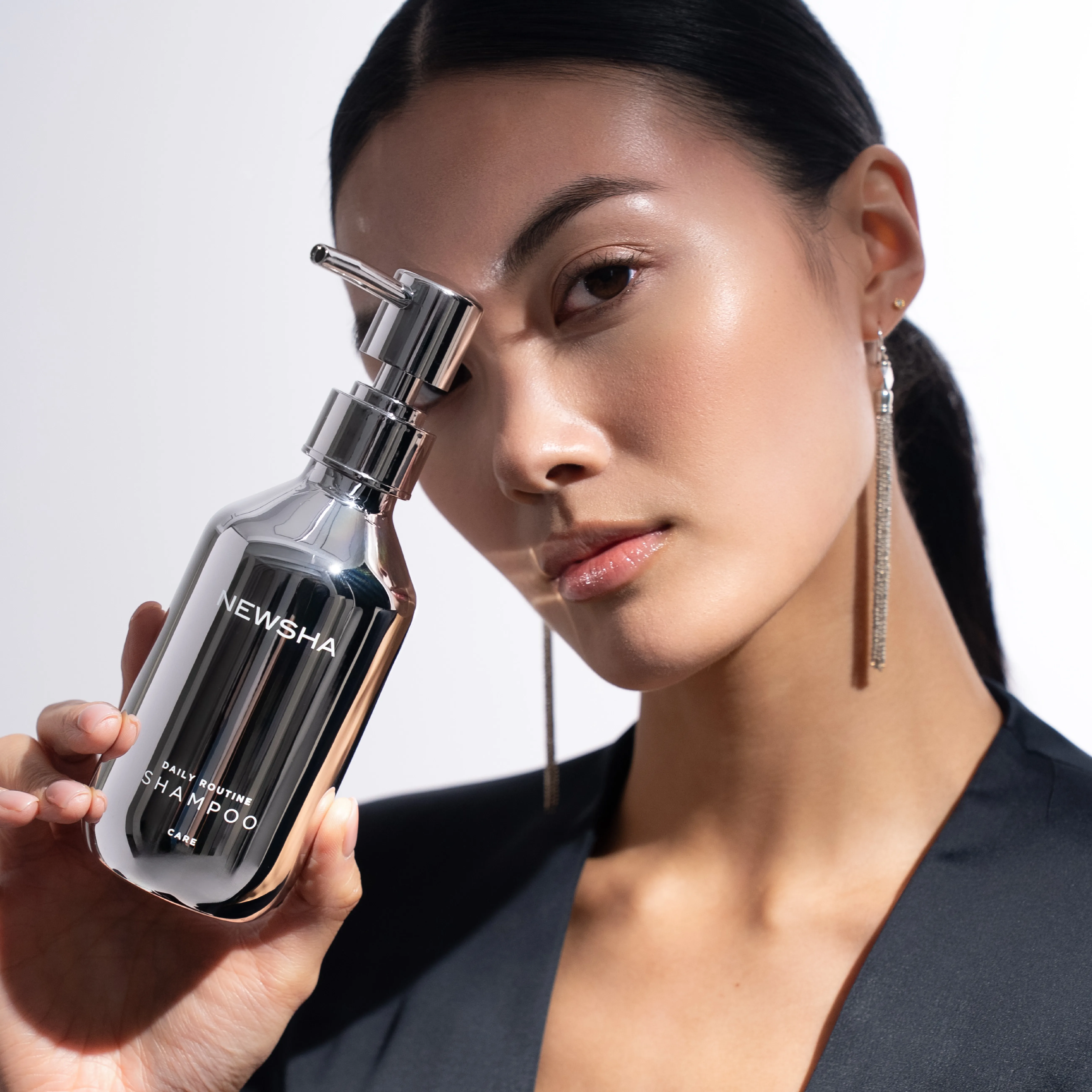 Frau mit sleekem Pferdeschwanz und schwarzem Blazer h&auml;lt eine silberne NEWSHA-Shampooflasche neben ihr Gesicht. Das minimalistische Beauty-Portr&auml;t setzt Produkt und gepflegte Ausstrahlung in den Fokus.