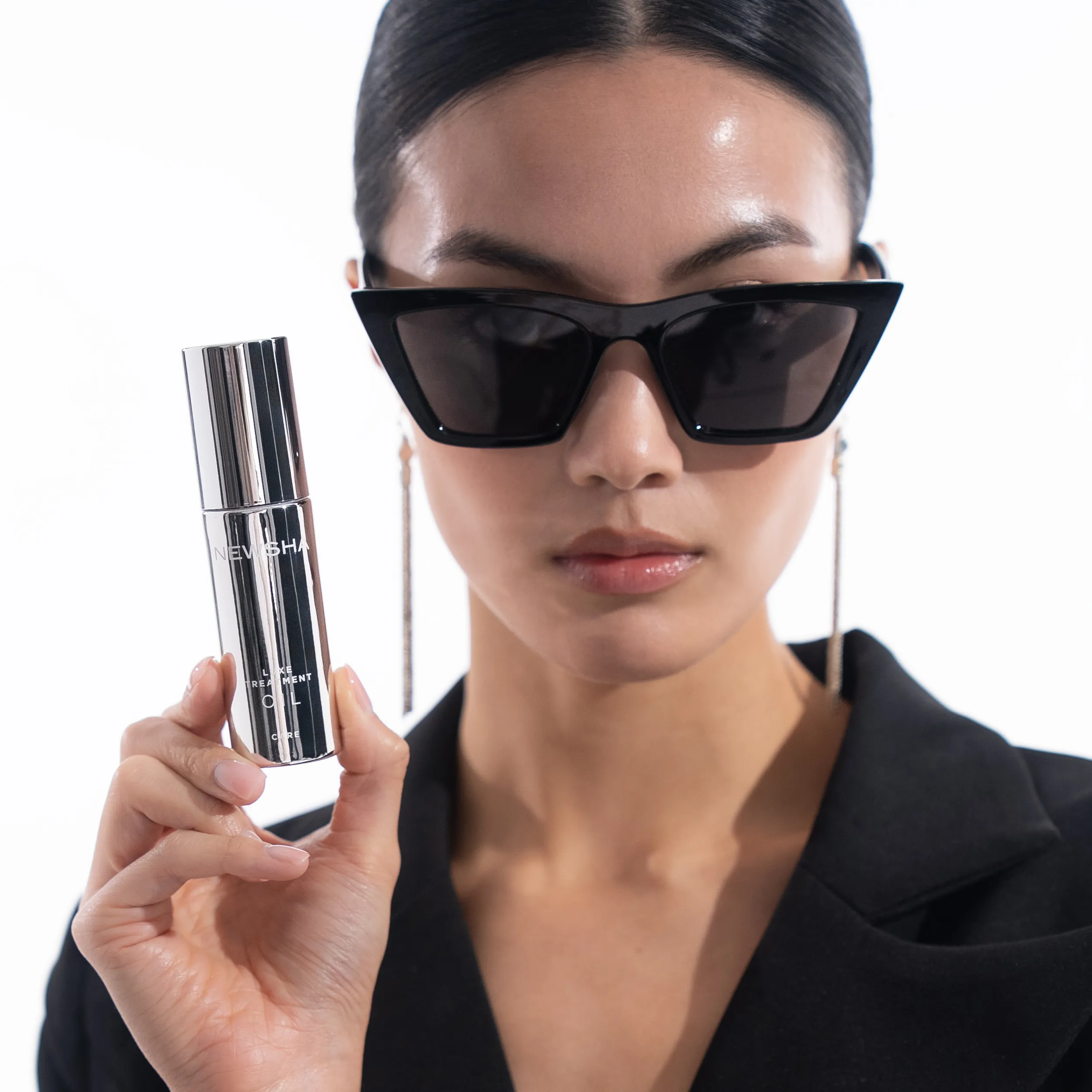 Model mit schwarzem Blazer, Cat-Eye-Sonnenbrille und langen Ohrringen h&auml;lt das silberne NEWSHA Luxe Treatment Oil vor hellem Studiohintergrund in die Kamera