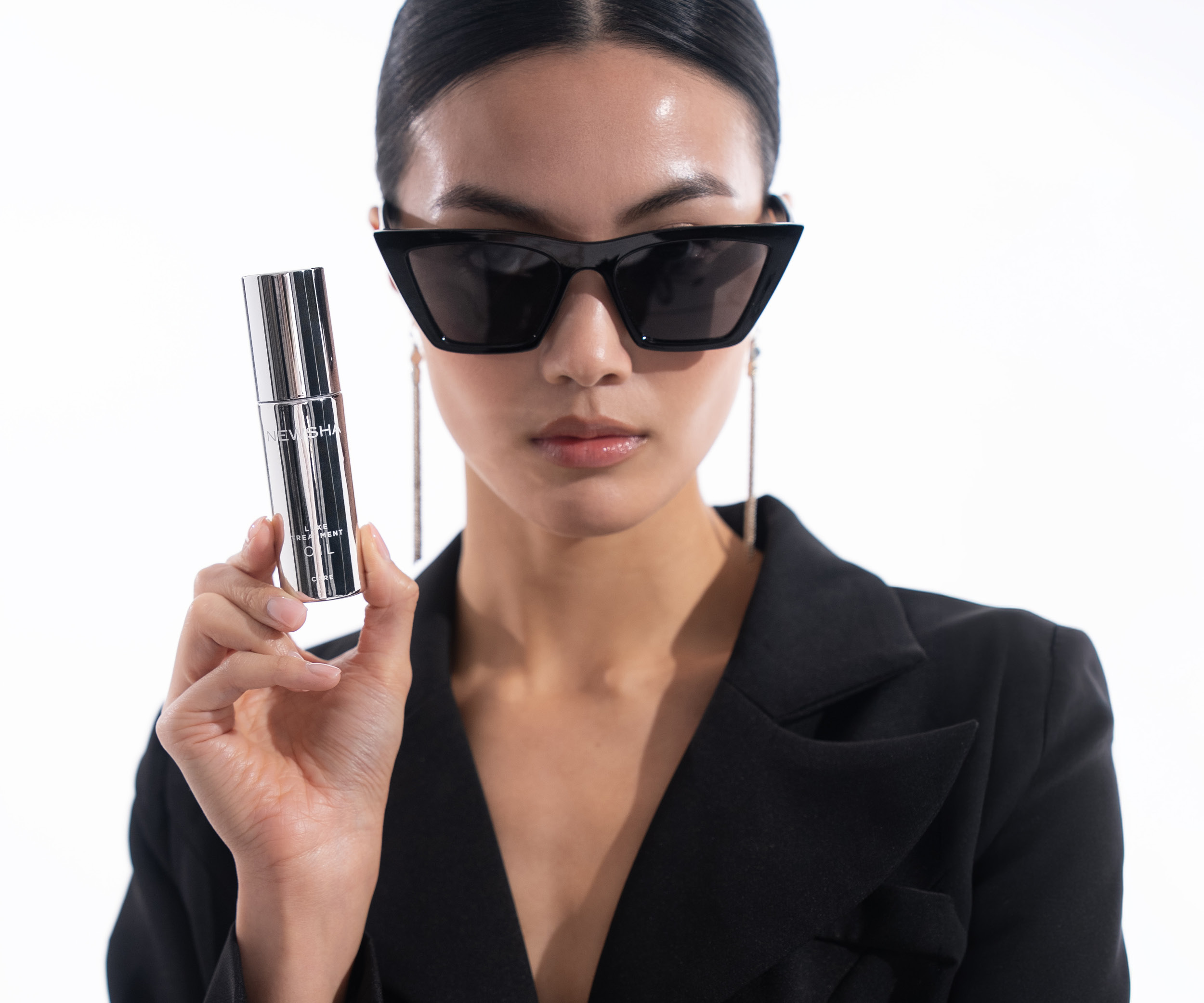 Model mit schwarzem Blazer, Cat-Eye-Sonnenbrille und langen Ohrringen h&auml;lt das silberne NEWSHA Luxe Treatment Oil vor hellem Studiohintergrund in die Kamera.