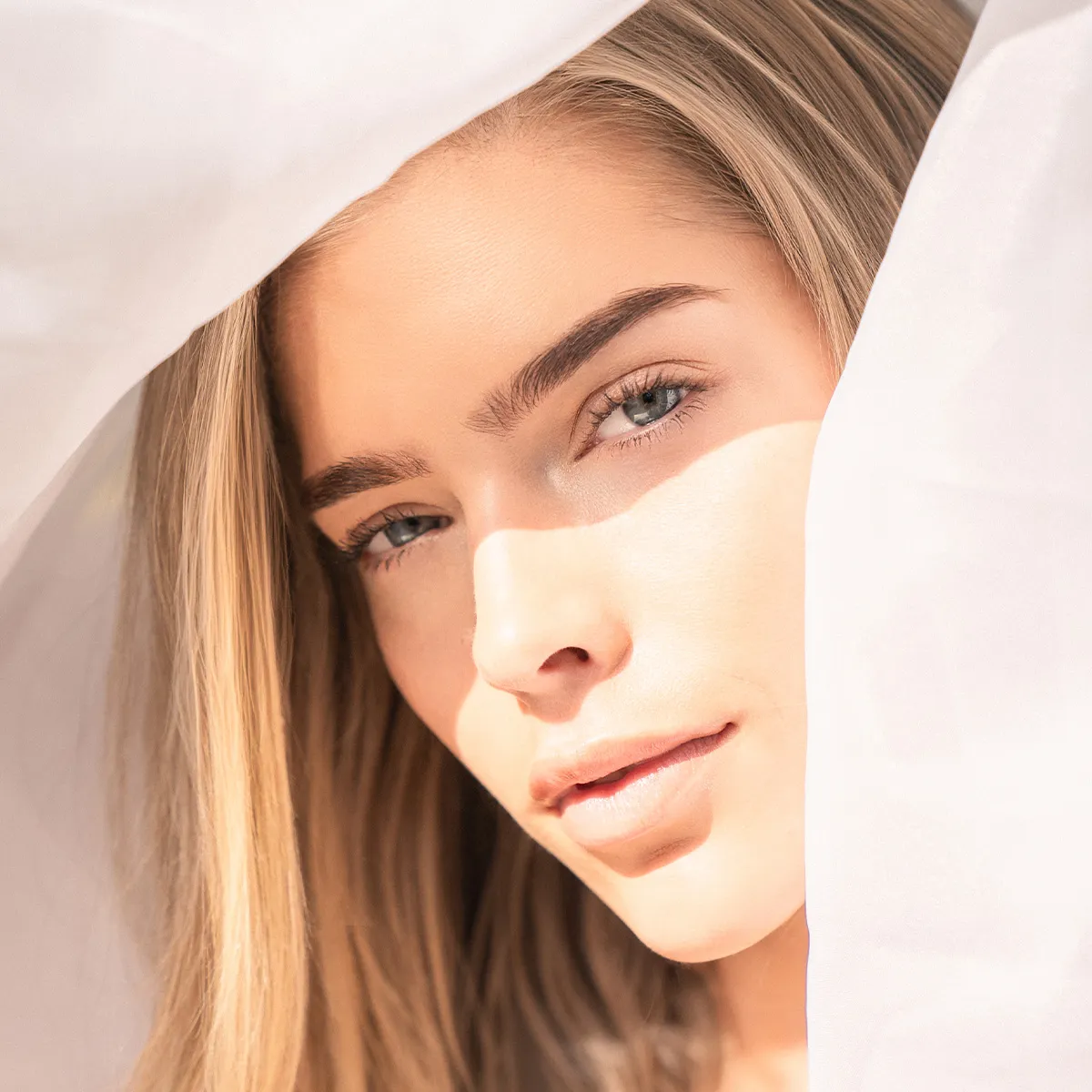 ahaufnahme eines blonden Models mit nat&uuml;rlichem Glow-Look, umrahmt von hellem Stoff und sanftem Sonnenlicht f&uuml;r eine weiche, elegante Beauty-Inszenierung.